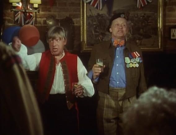 Eric in a red waistcoat and Tinker in uniform and beret, medals and drink in hand, under Union Jack bunting at the opening night party