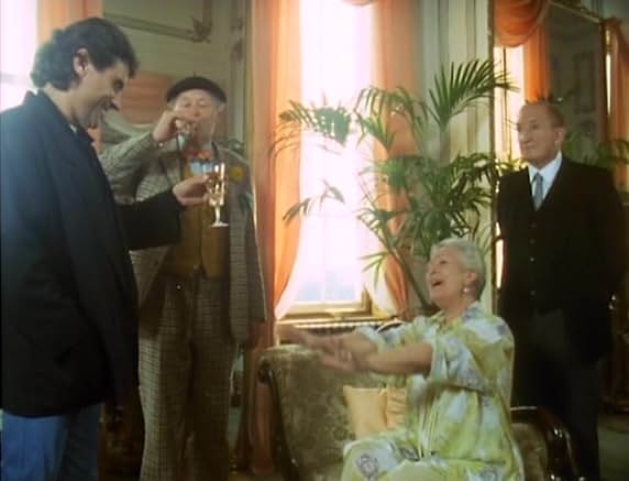 Madelene Gilbert seated in her drawing room raising a hand to Lovejoy and Tinker, who is drinking champagne, with her butler Michael standing by