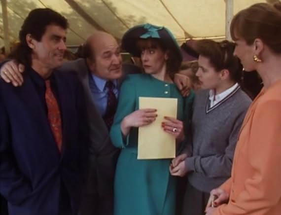 Lovejoy in a navy suit and orange tie in the parents' day marquee, wedged beside a bald man in a grey suit, with Susan in a teal hat and suit holding a yellow envelope, Vicky in school uniform and another guest in peach