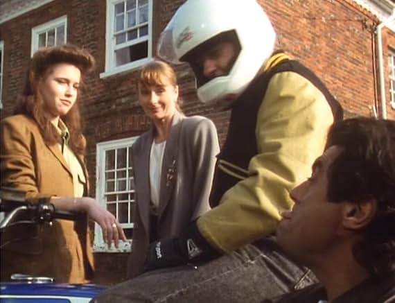 Susan and Vicky outside a red-brick building, one in a tan jacket and one in a grey cardigan, with Eric between them on his motorbike in his white Lazer crash helmet, and Lovejoy in the foreground looking up from a car